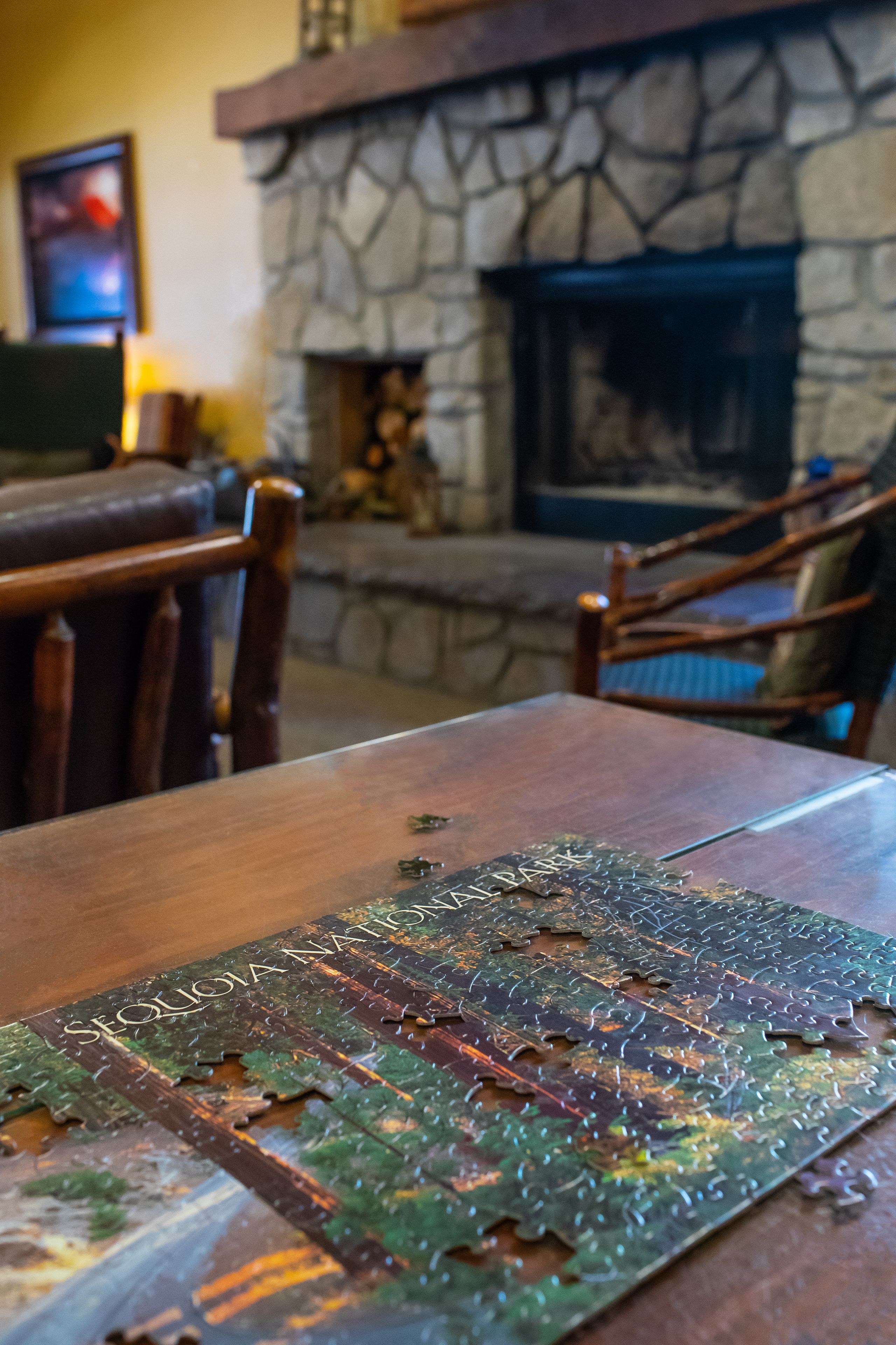 A wooden table with a partially completed jigsaw puzzle featuring a forest scene. In the background, there is a stone fireplace with a fire, and wooden chairs with cushions.