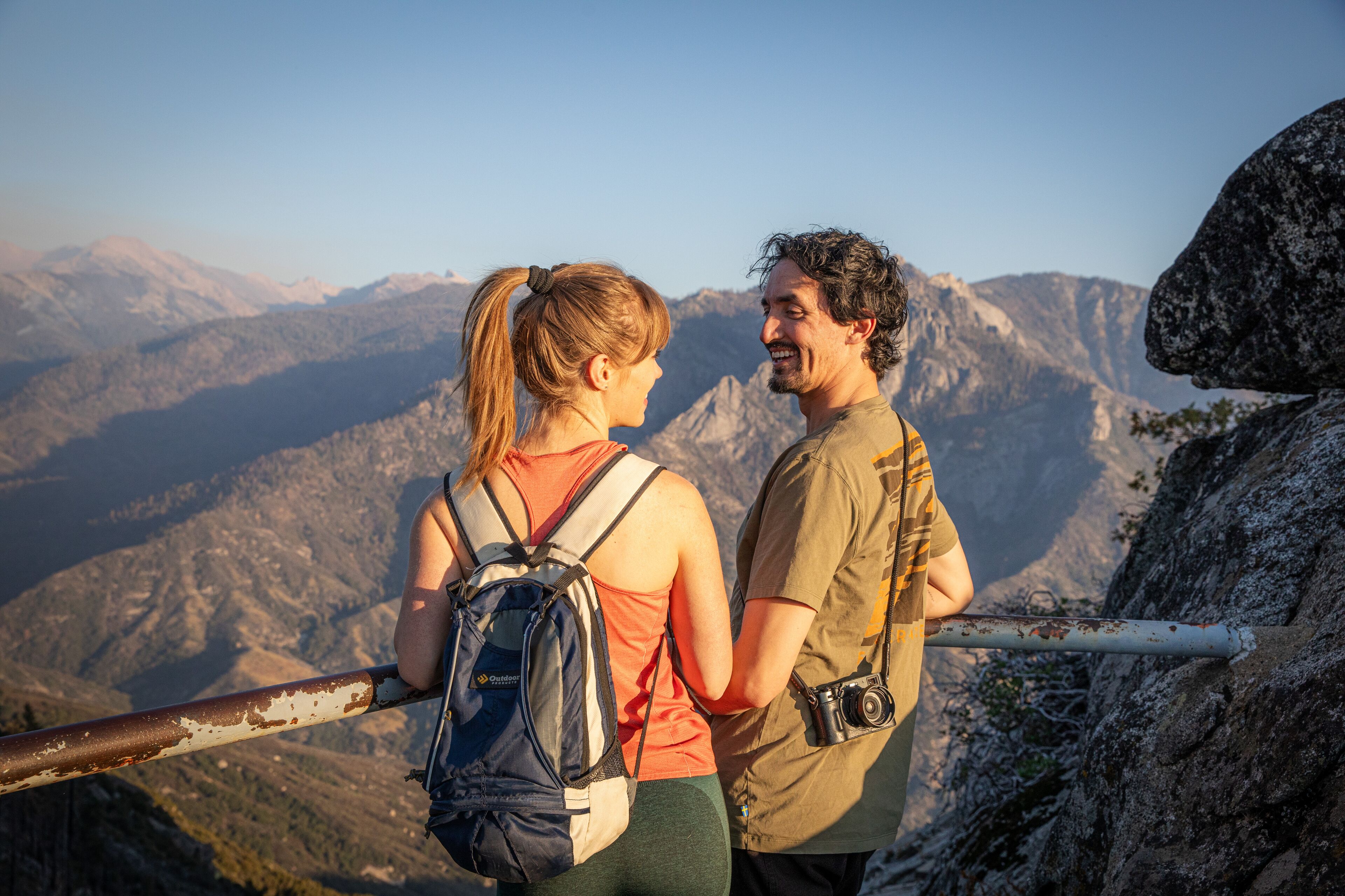 Two people standing on a mountain trail with a rocky path and a metal railing.
