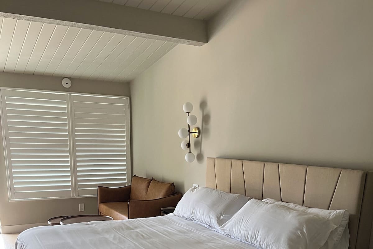 A neatly made bed in a room with a sloped ceiling, a wall-mounted light fixture, and a window with blinds.
