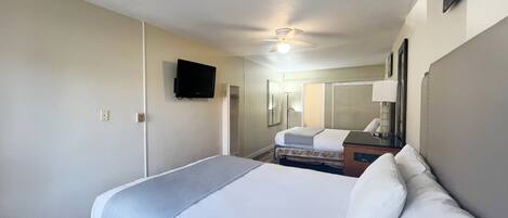 A hotel room with a bed, a television mounted on the wall, a ceiling fan, and a window with blinds.