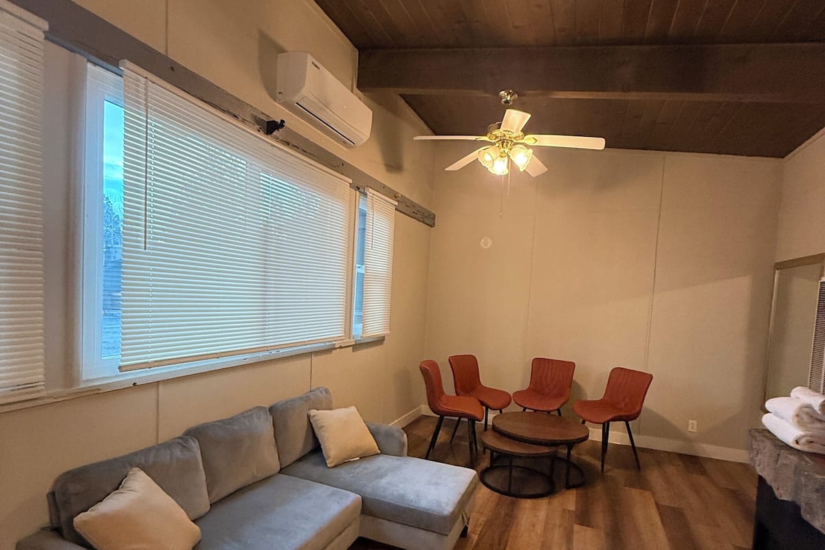 A modern living room with a gray sectional sofa, a wooden ceiling, and a dining area with red chairs and a round table.