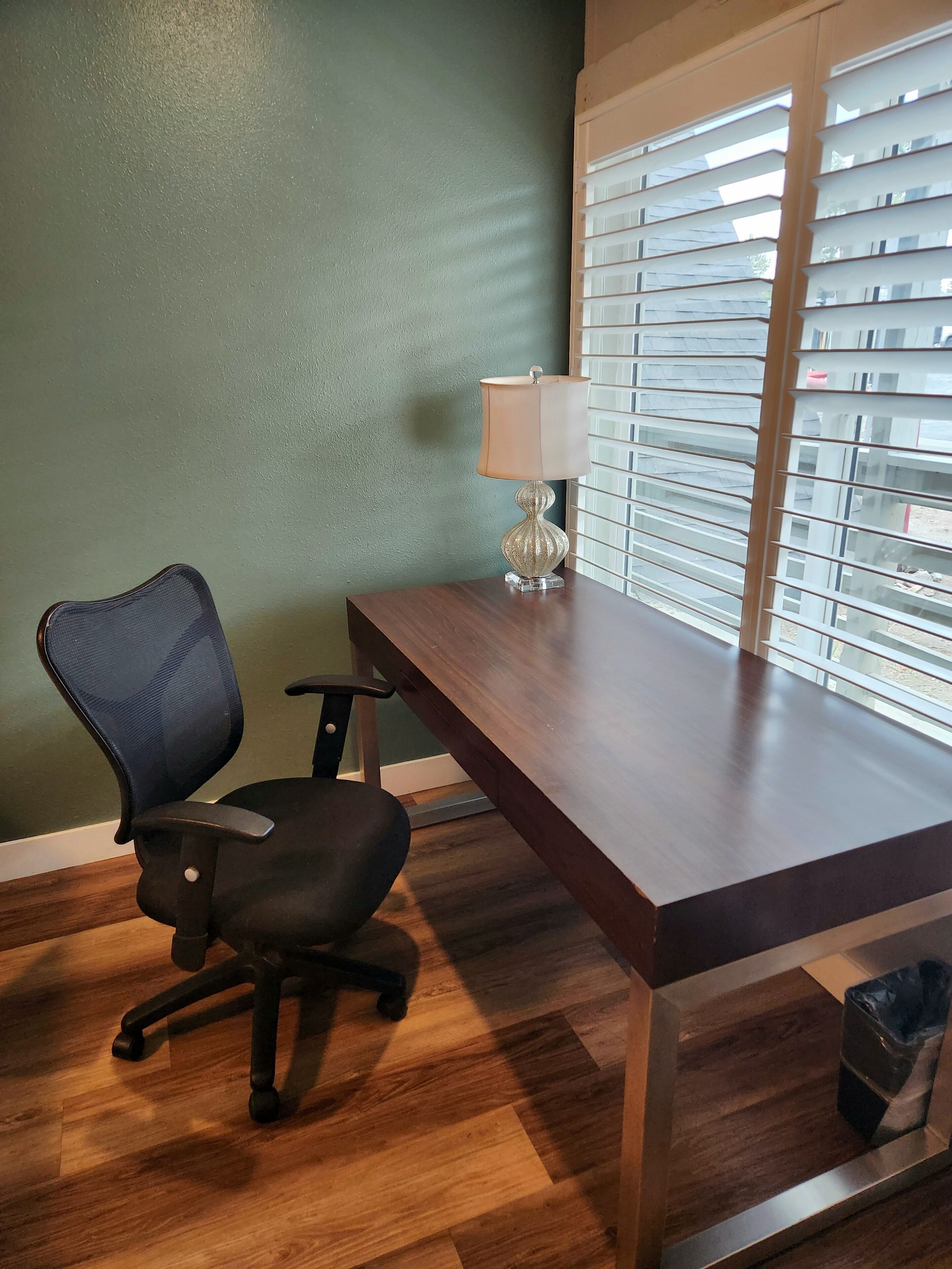 A wooden desk with a lamp, a black office chair, and a window with blinds.
