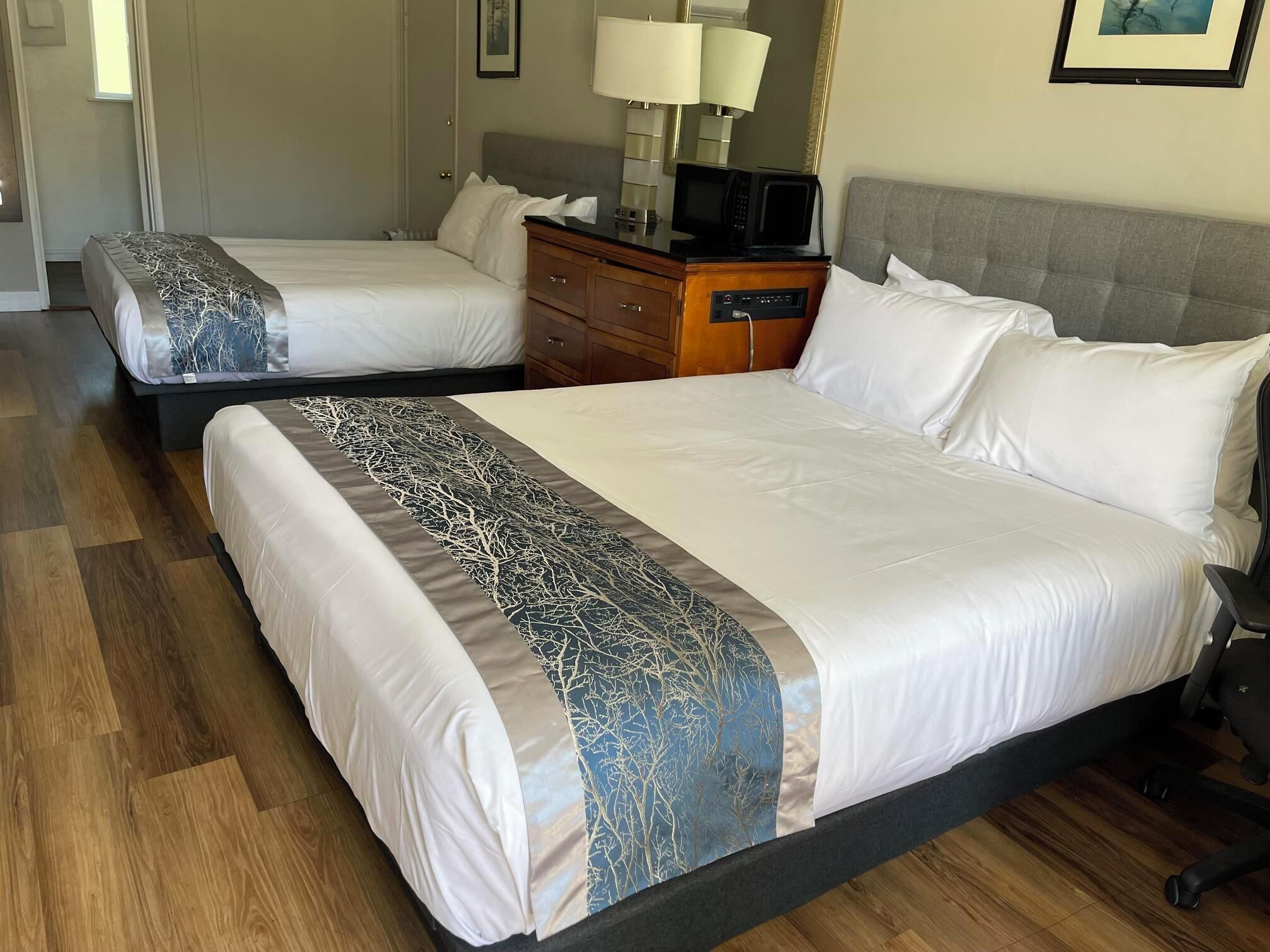 A hotel room with two beds, wooden flooring, a dresser with a lamp, and a microwave on the dresser.