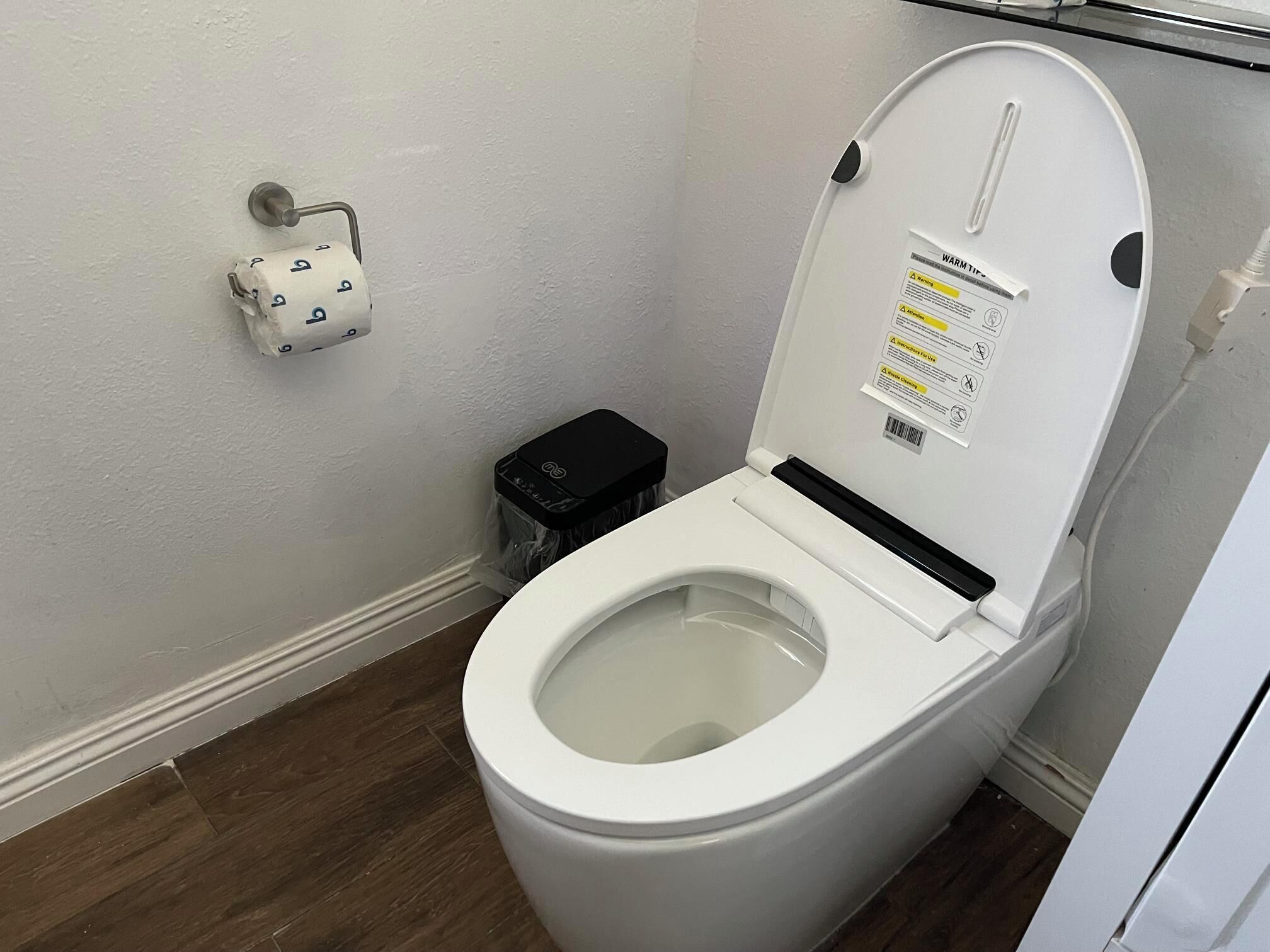 Een wit toilet met gesloten deksel, een toiletrolhouder aan de muur en een zwarte prullenbak.