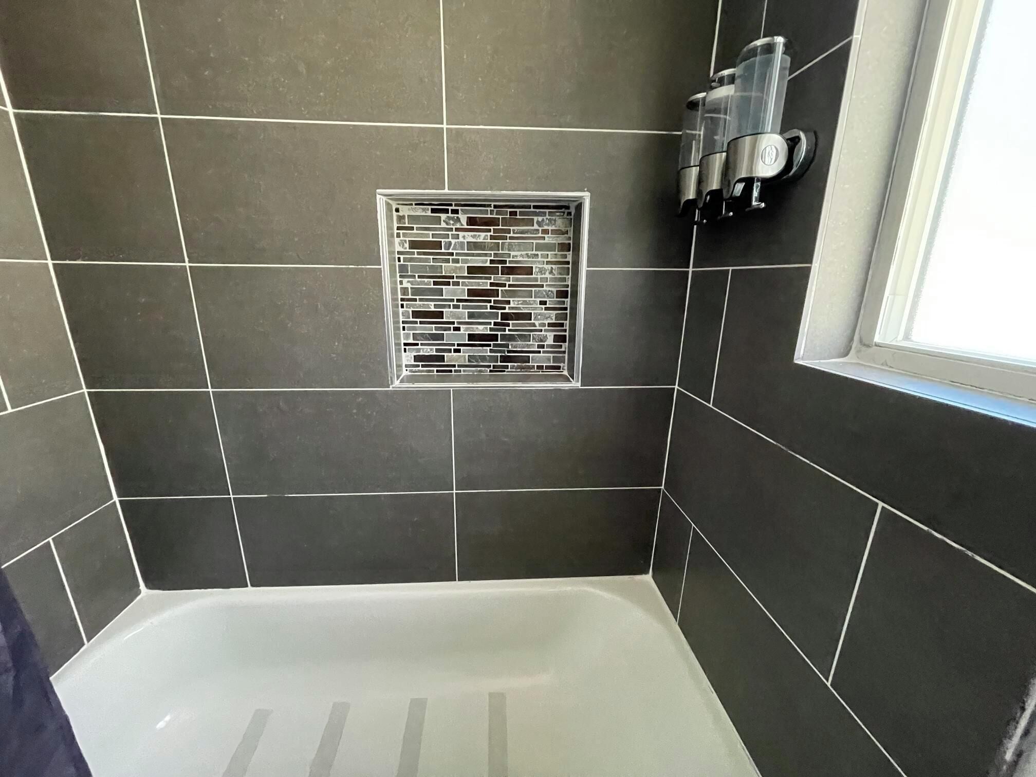 Een badkamer met donkere tegels, een witte badkuip, een douchegedeelte en een raam.