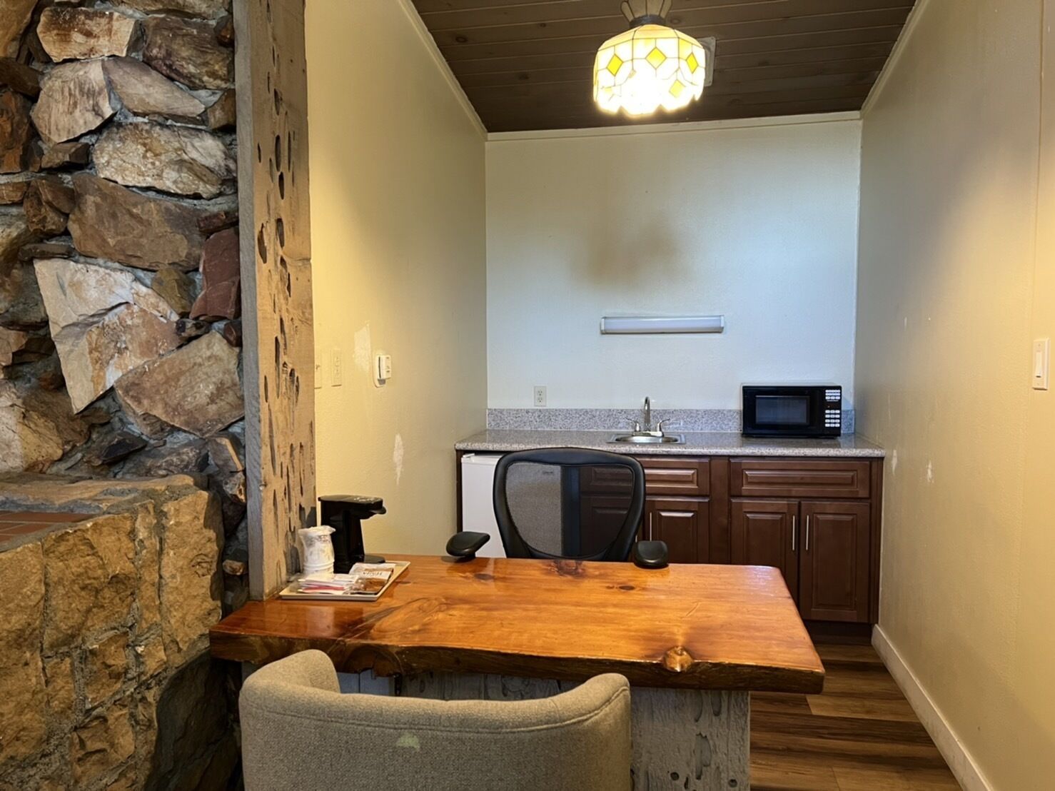 Uma cozinha compacta com parede de pedra decorativa, uma mesa de jantar de madeira, uma cadeira de escritório preta e um micro-ondas.
