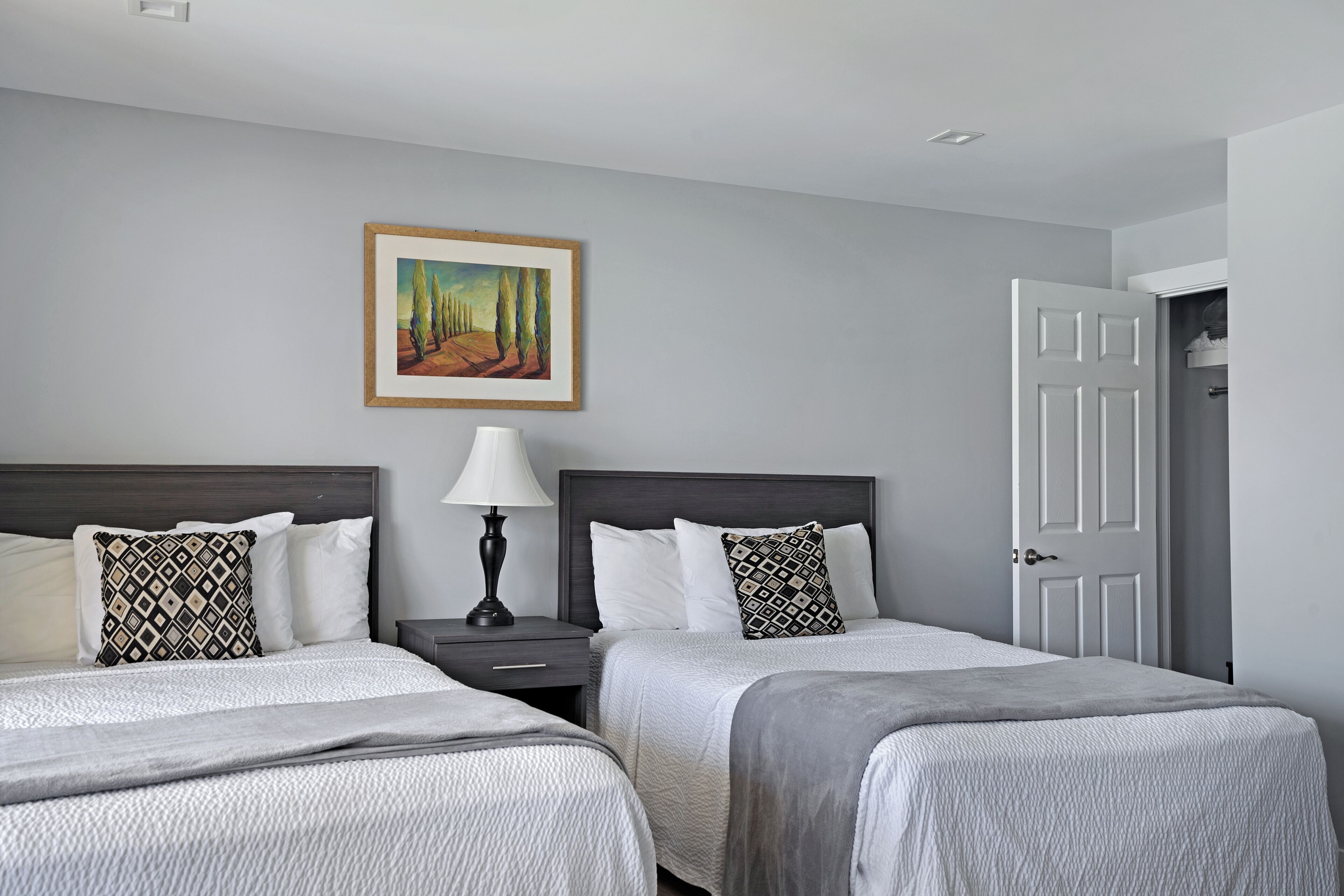 A hotel room with two beds, a nightstand, a lamp, a painting on the wall, and a door.