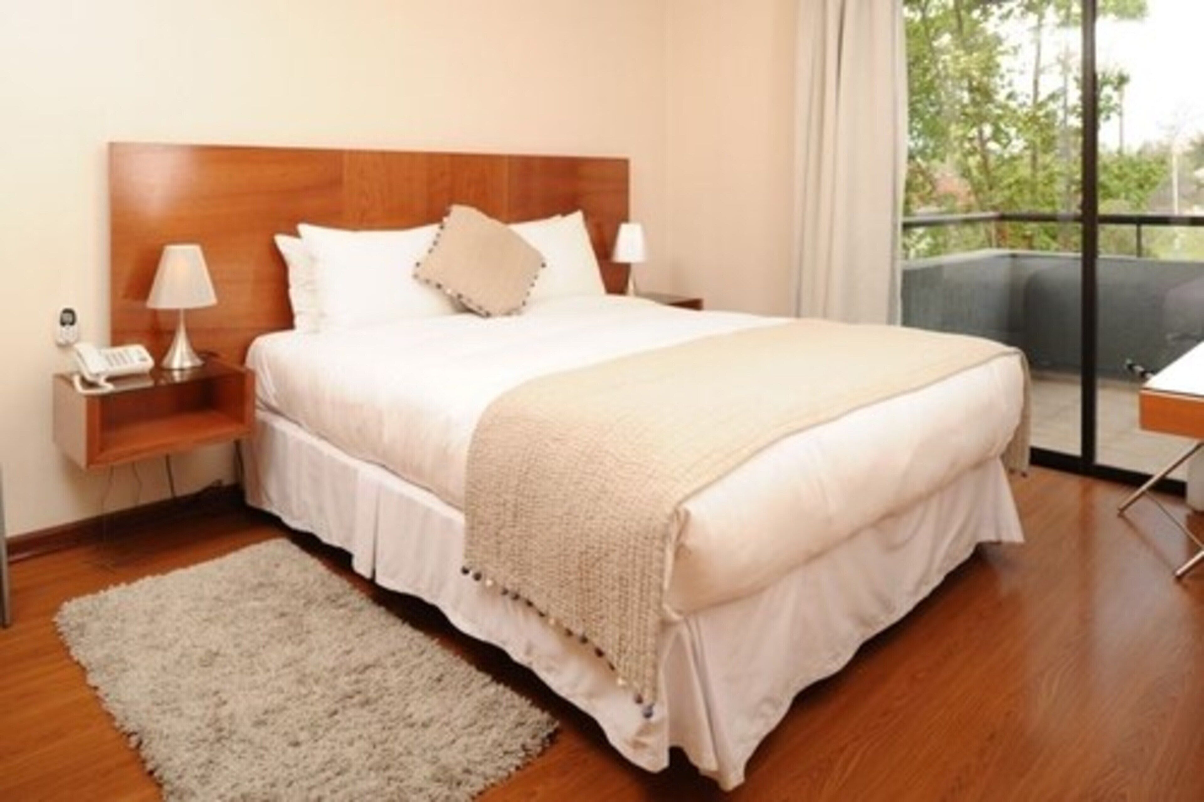 A neatly made bed with a wooden headboard, a bedside table with a lamp, and a view of the outdoors through a balcony.