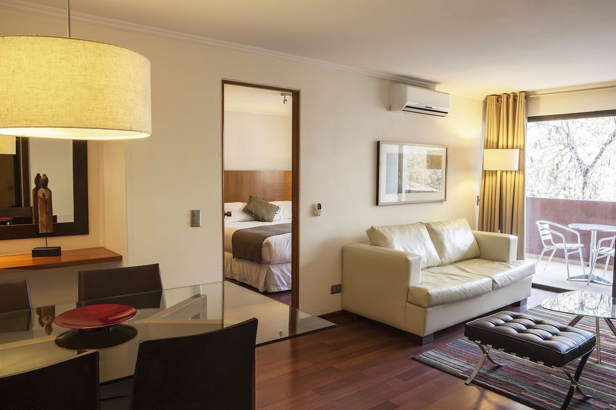 Una habitación de hotel moderna con una mesa de comedor de cristal, un sofá, una banca acolchada, un espejo grande y vistas al exterior.