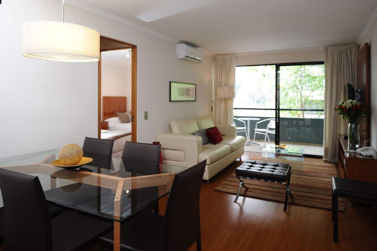 Una sala de estar moderna con una mesa de vidrio, un sofá y una mesa de centro. Se aprecia un ventanal con cortinas, un ventilador de techo y un cuadro en la pared.