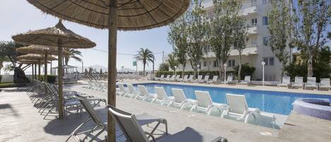 A poolside area with lounge chairs and thatched umbrellas, a swimming pool, and a building with balconies.