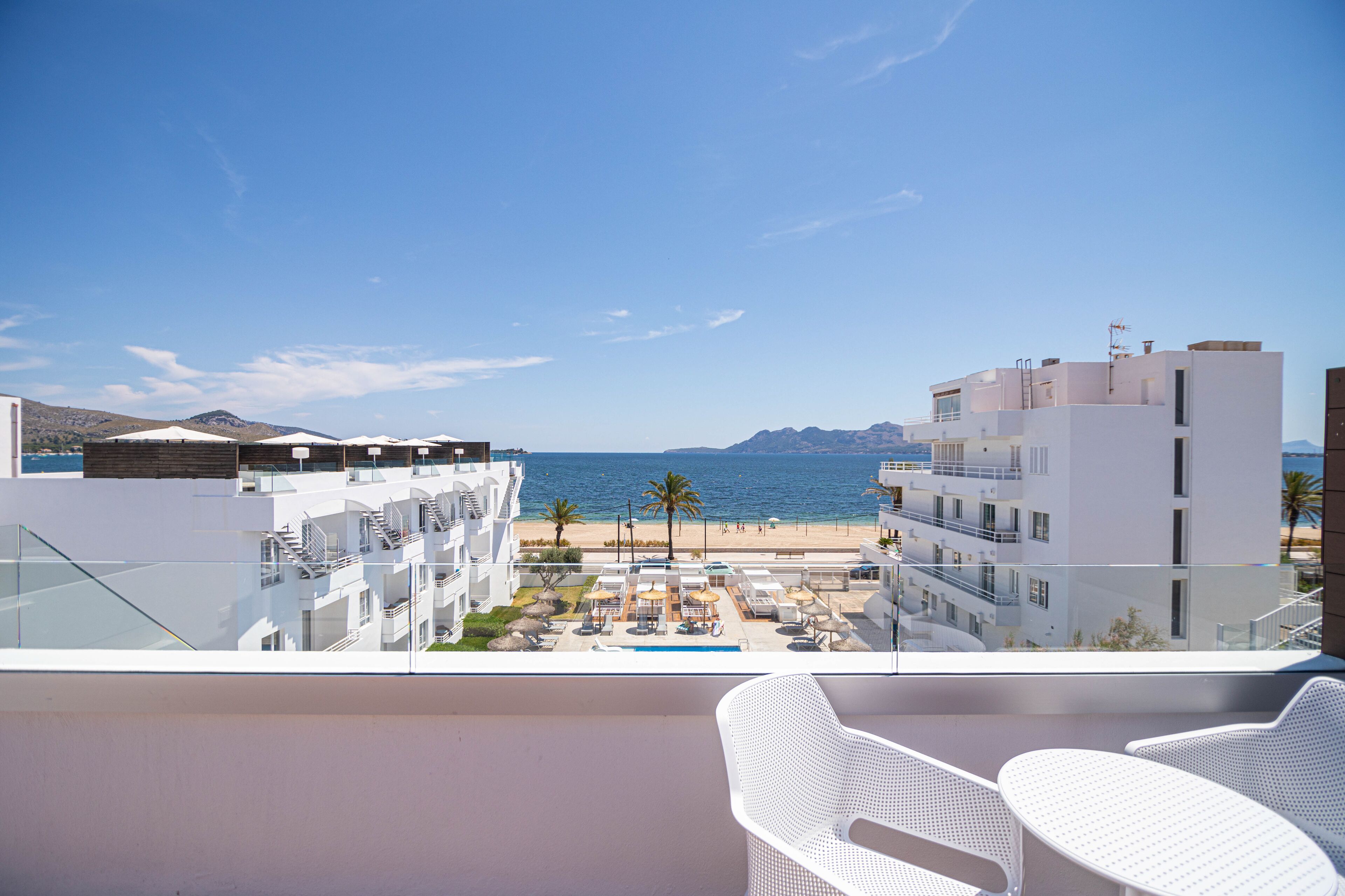 A balcony with white wicker chairs, overlooking a pool area with umbrellas, a beach, and a mountain range.