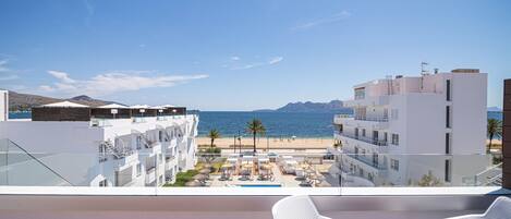 A balcony with white wicker chairs, overlooking a pool area with umbrellas, a beach, and a mountain range.