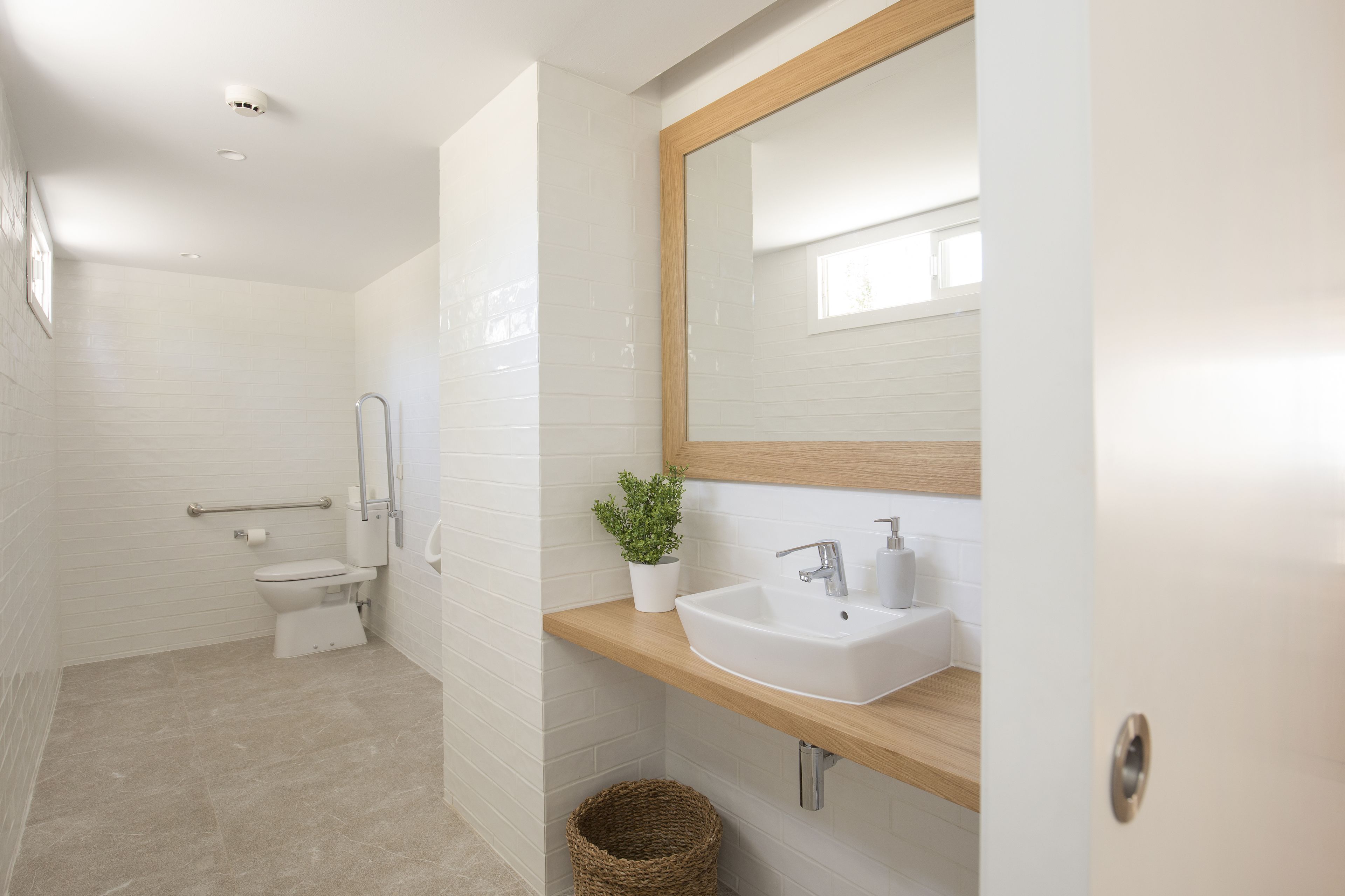 Un baño moderno con azulejos blancos, tocador de madera, lavamanos, espejo y retrete.