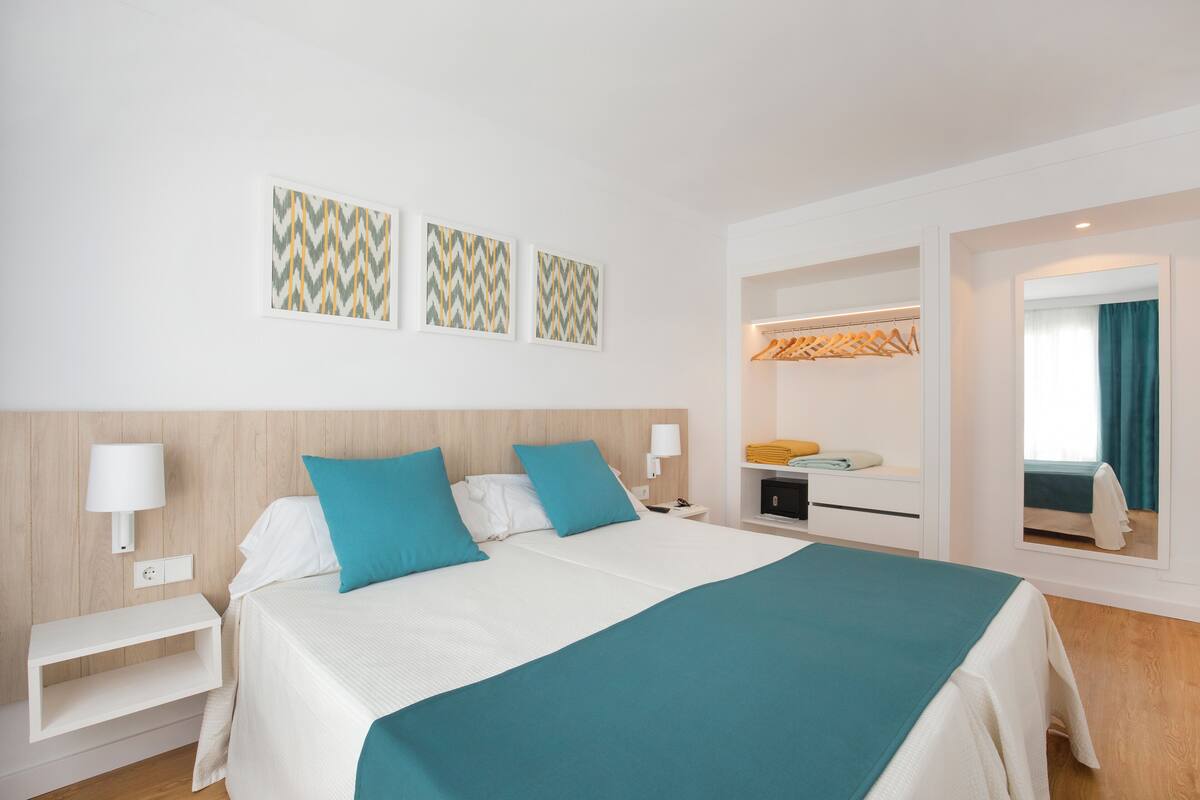 A hotel room with a large bed, two pillows, a bedside table, a nightstand, a mirror, and a wardrobe.