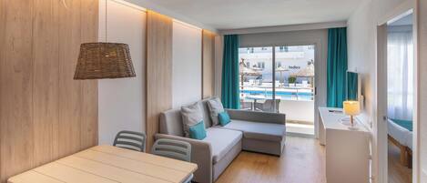 Une chambre d’hôtel moderne avec un mur d’accent en bois, un coin repas avec une table et des chaises, un canapé avec des coussins turquoise et une vue sur la piscine et les bâtiments environnants.