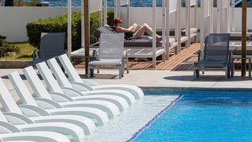 A pool area with lounge chairs and umbrellas, a person relaxing, and a view of the sea.