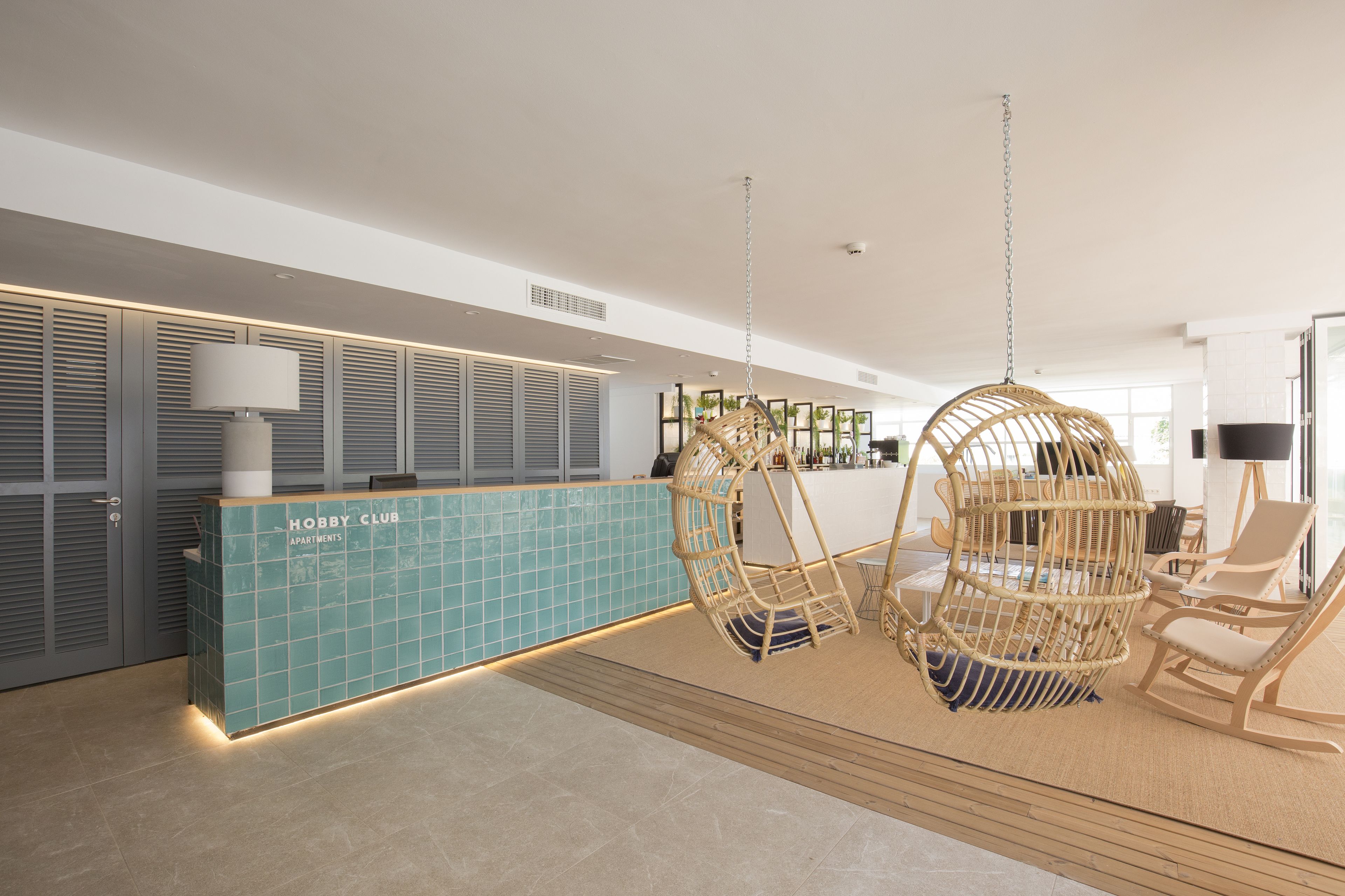 A reception area with a blue tiled counter labeled "Hobby Club Apartments," wicker hanging chairs, and a wooden floor.