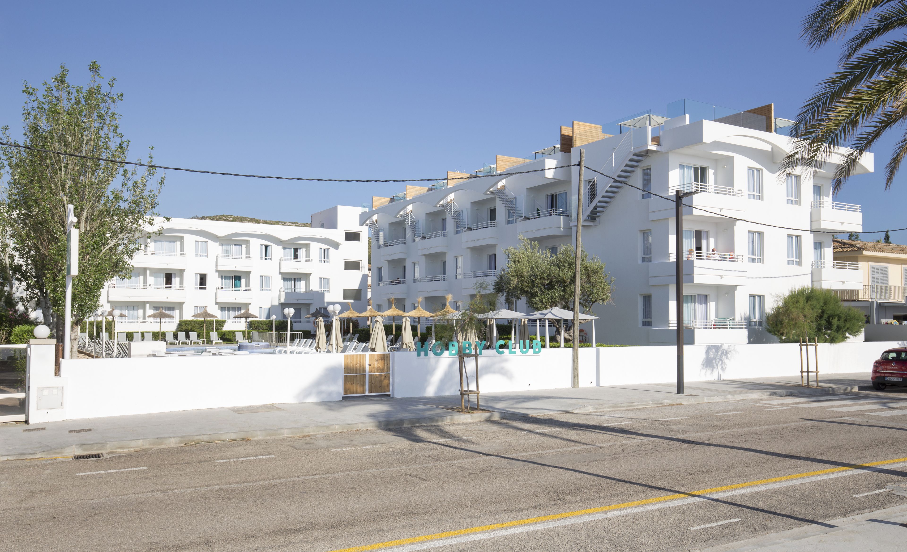 Un bâtiment moderne de couleur blanche, doté de nombreux balcons, un espace piscine avec des chaises longues et un ciel d’un bleu éclatant.
