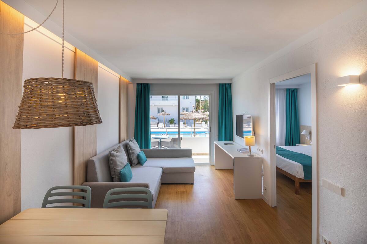 A modern hotel room with a dining area, a sofa, a desk, and a view of the pool.