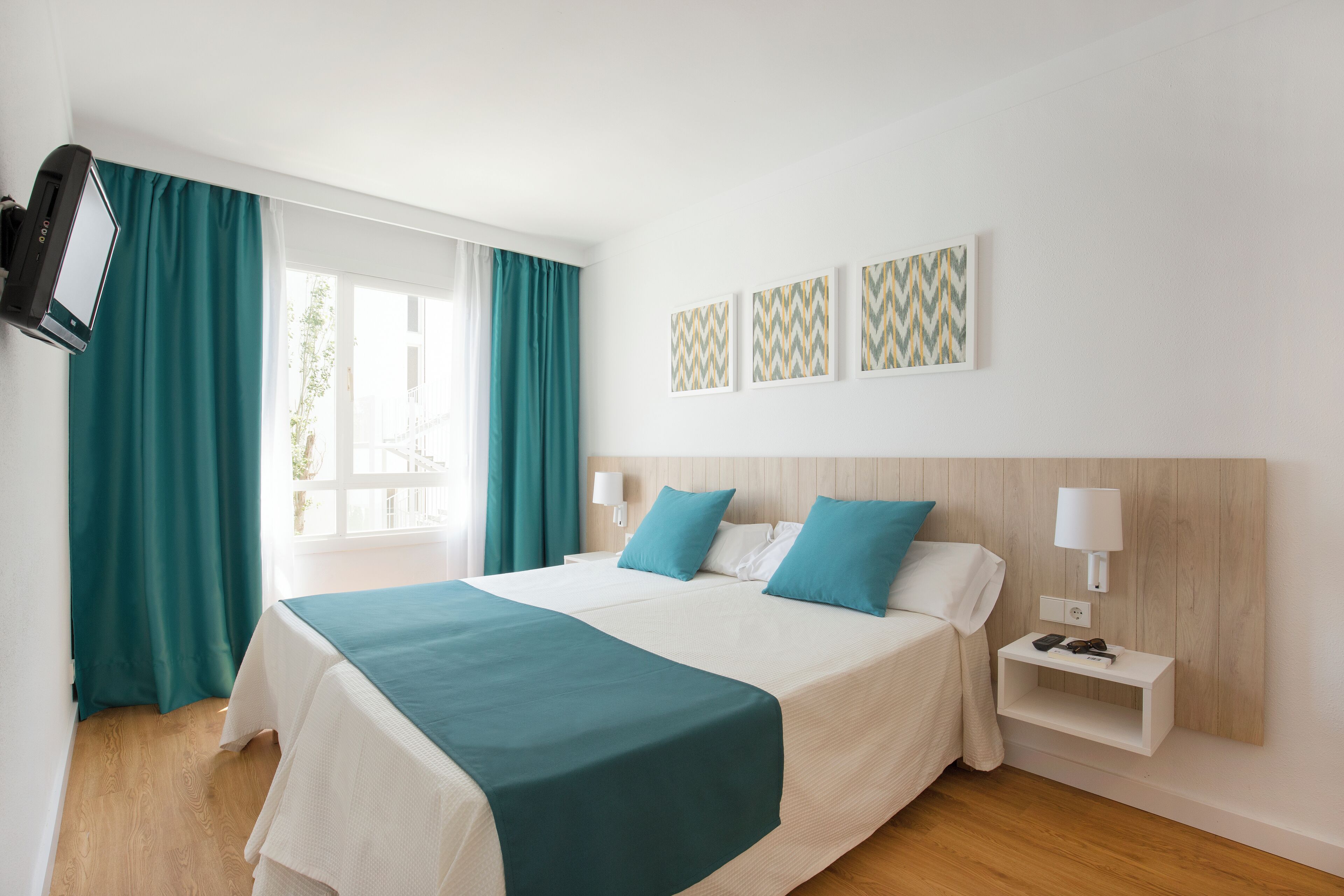 Habitación de hotel con cama, cortinas verde azulado, televisor y tres piezas de arte mural estampadas.