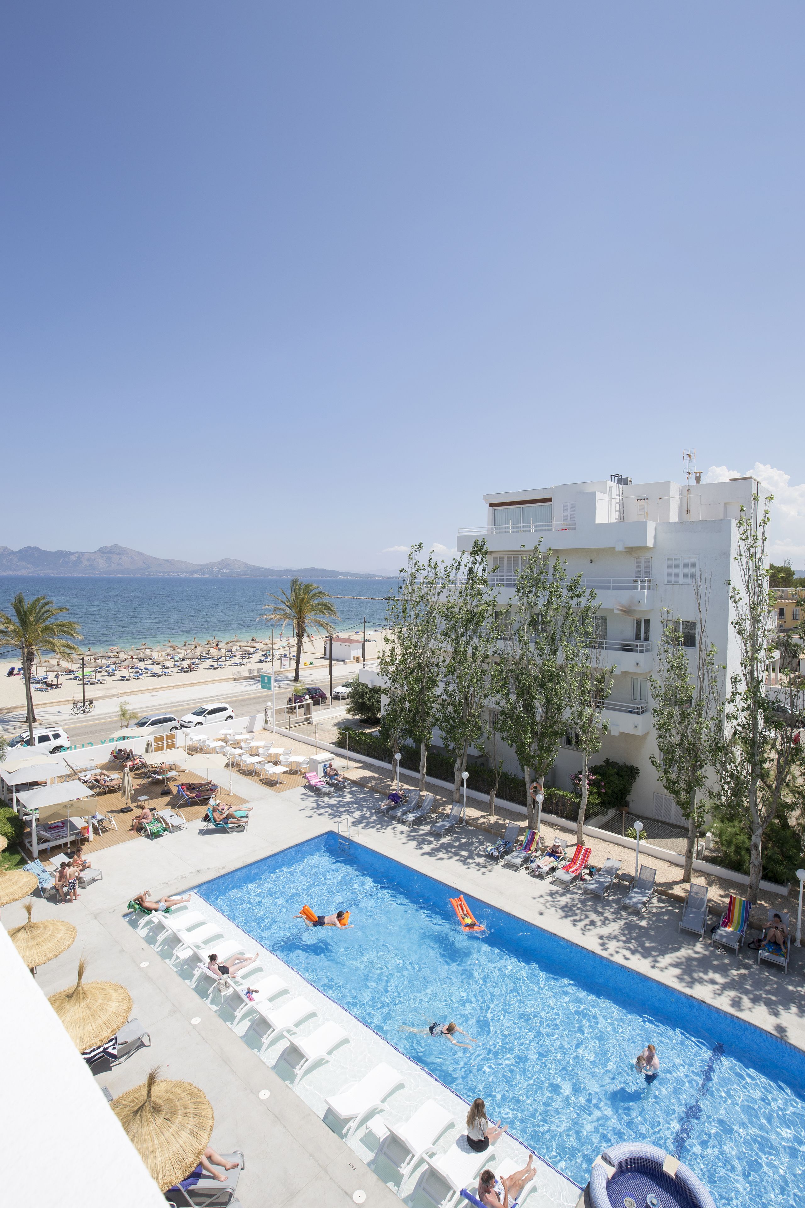 A pool area with lounge chairs and umbrellas, a building with balconies, and a view of the sea.