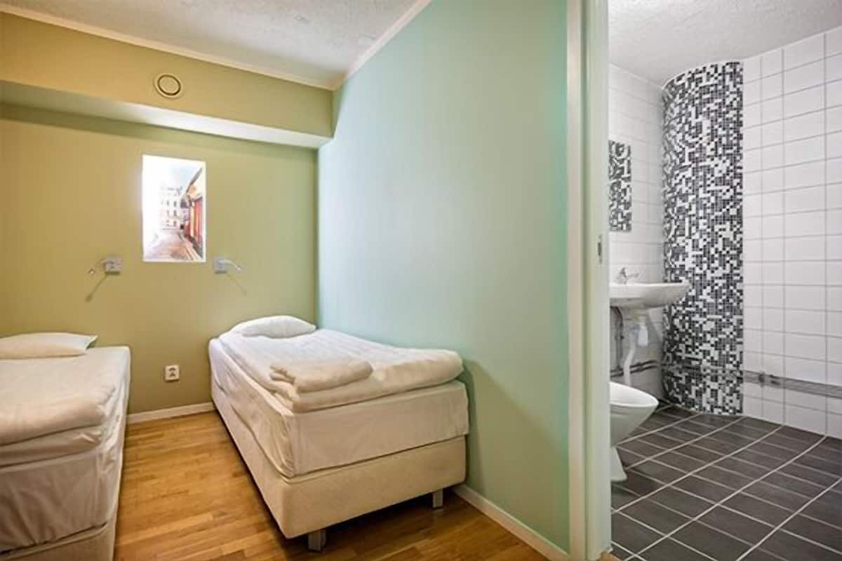 A small, single-room apartment with a bed, a window, and a bathroom with a tiled floor and a sink.