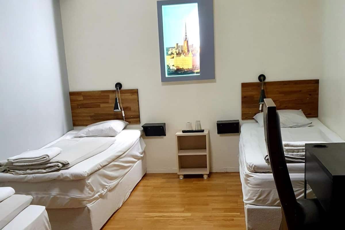A hotel room with two single beds, a desk, a chair, a small nightstand, and a framed picture on the wall.