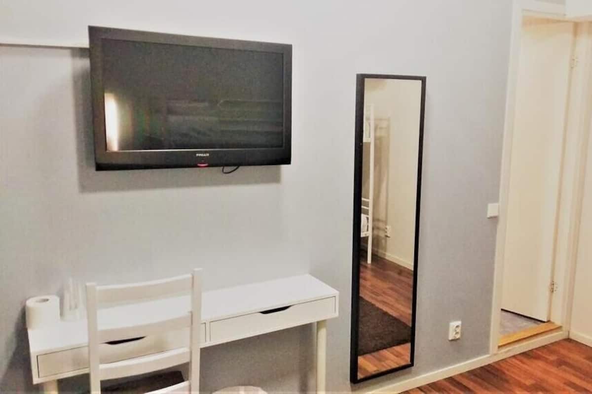 A room with a flat-screen TV mounted on the wall, a white desk with a chair, a large mirror, and a door.