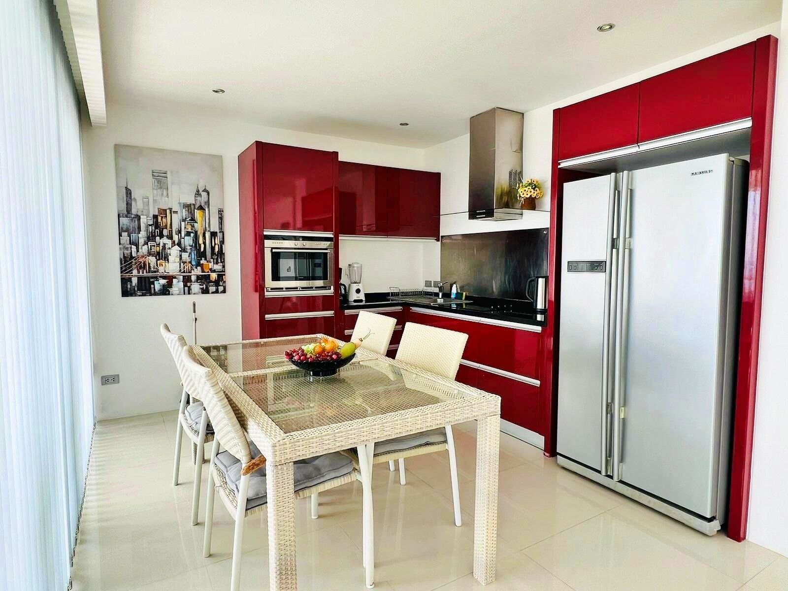 A modern kitchen with red cabinets, a stainless steel refrigerator, and a dining area with wicker chairs and a glass table.