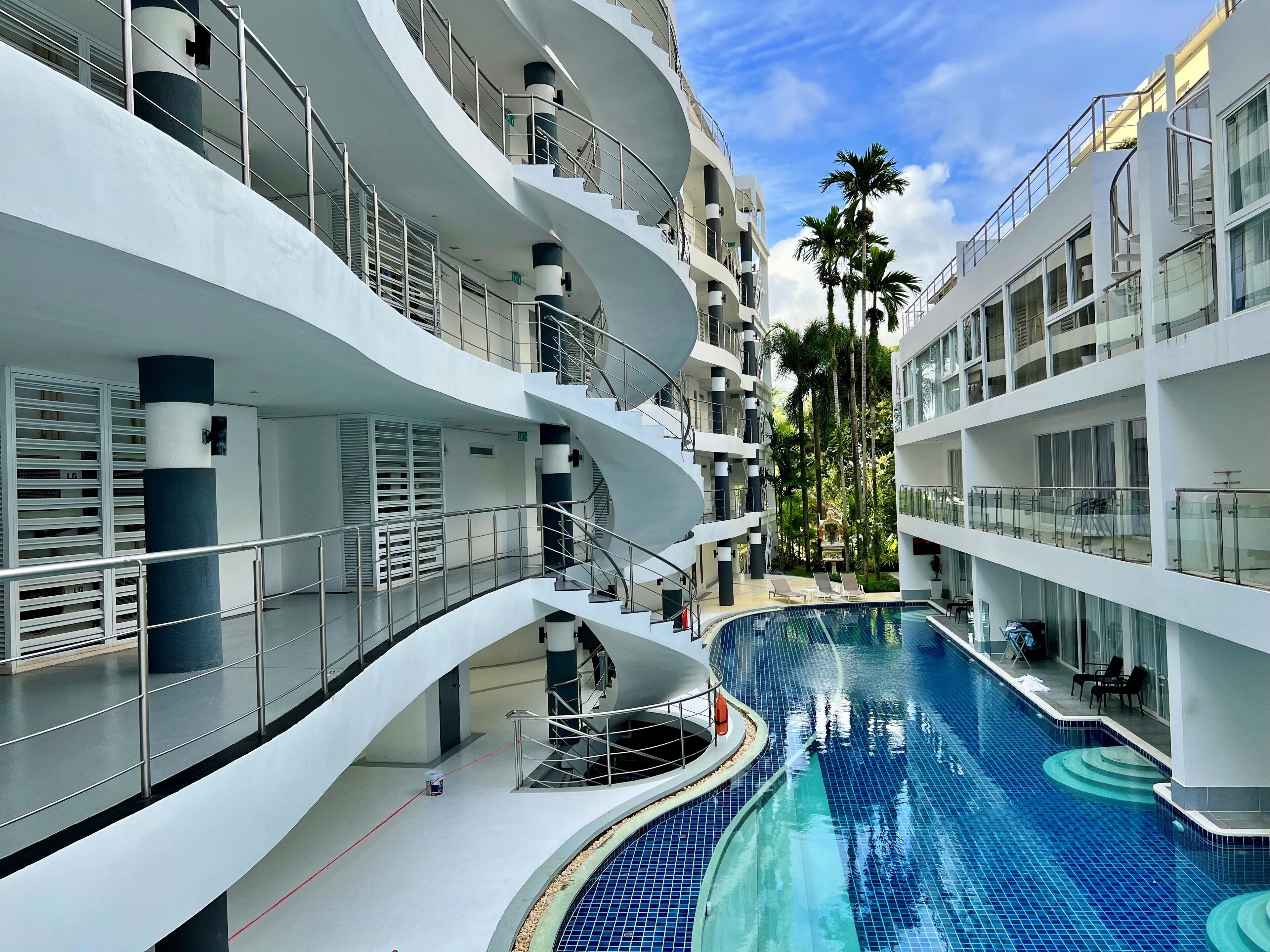 A modern multi-level building with a central swimming pool, balconies, and palm trees.