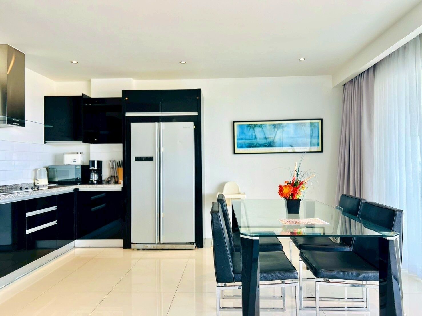 A modern kitchen with black cabinets, a stainless steel refrigerator, and a dining area with a glass table and black chairs.