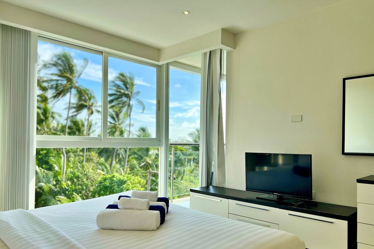 Una habitación de hotel moderna con una cama grande, un televisor de pantalla plana y vistas a palmeras a través de amplios ventanales.