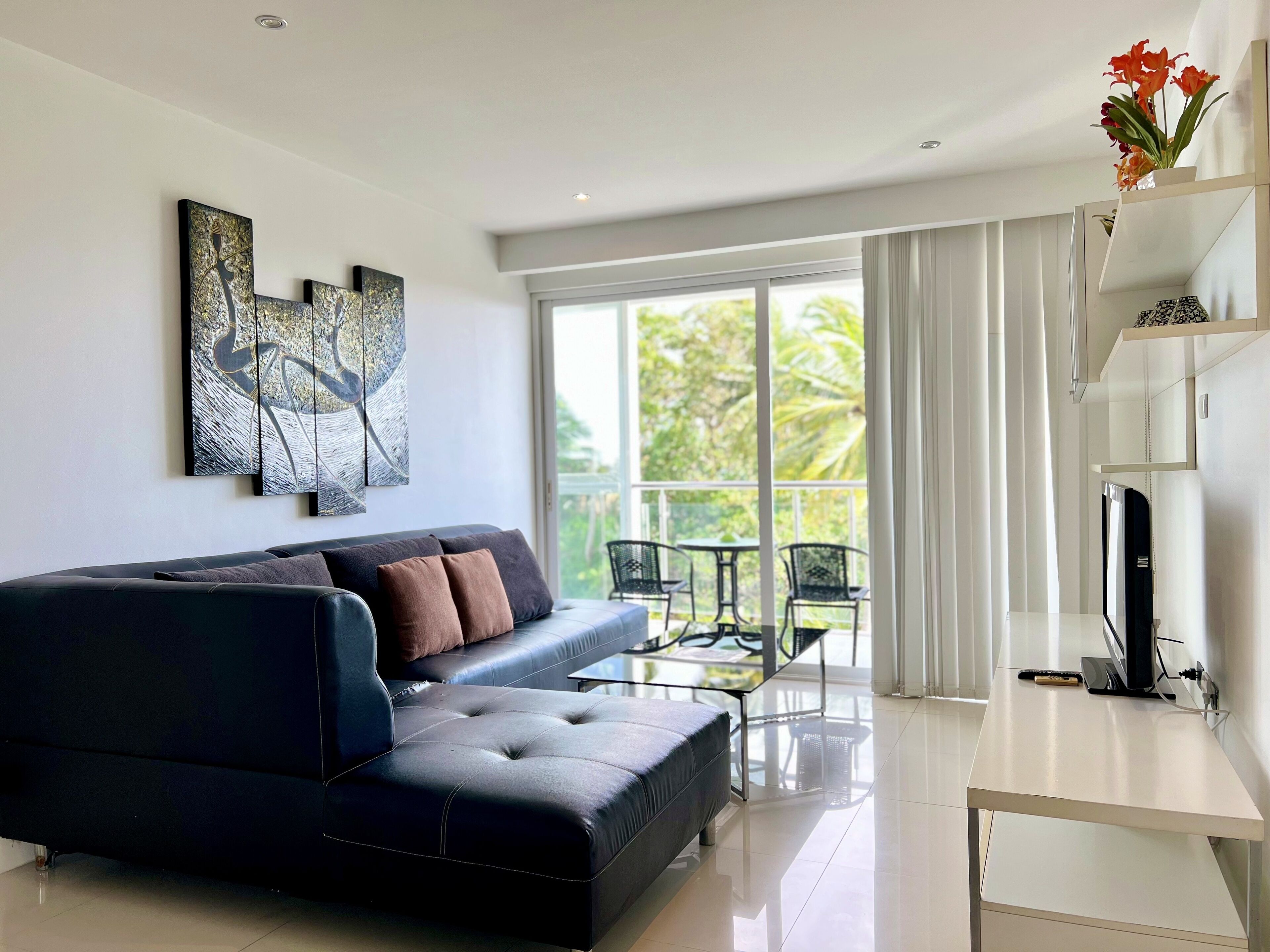 A modern living room with a black leather sofa, a glass coffee table, and a white TV stand. There are abstract paintings on the wall and a balcony with outdoor furniture.