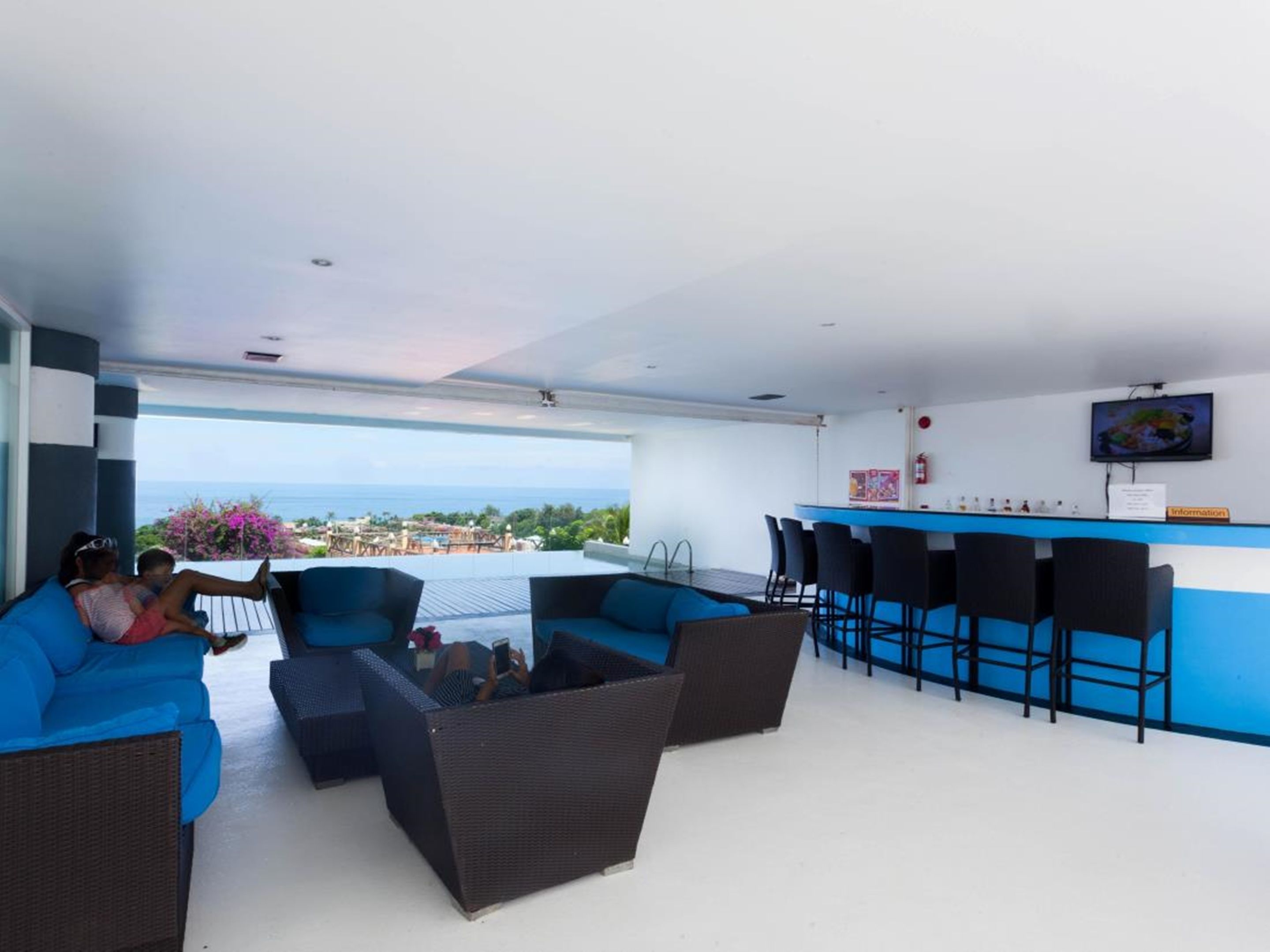 A modern lounge area with blue sofas and wicker chairs, a bar with black chairs, and a view of the ocean.