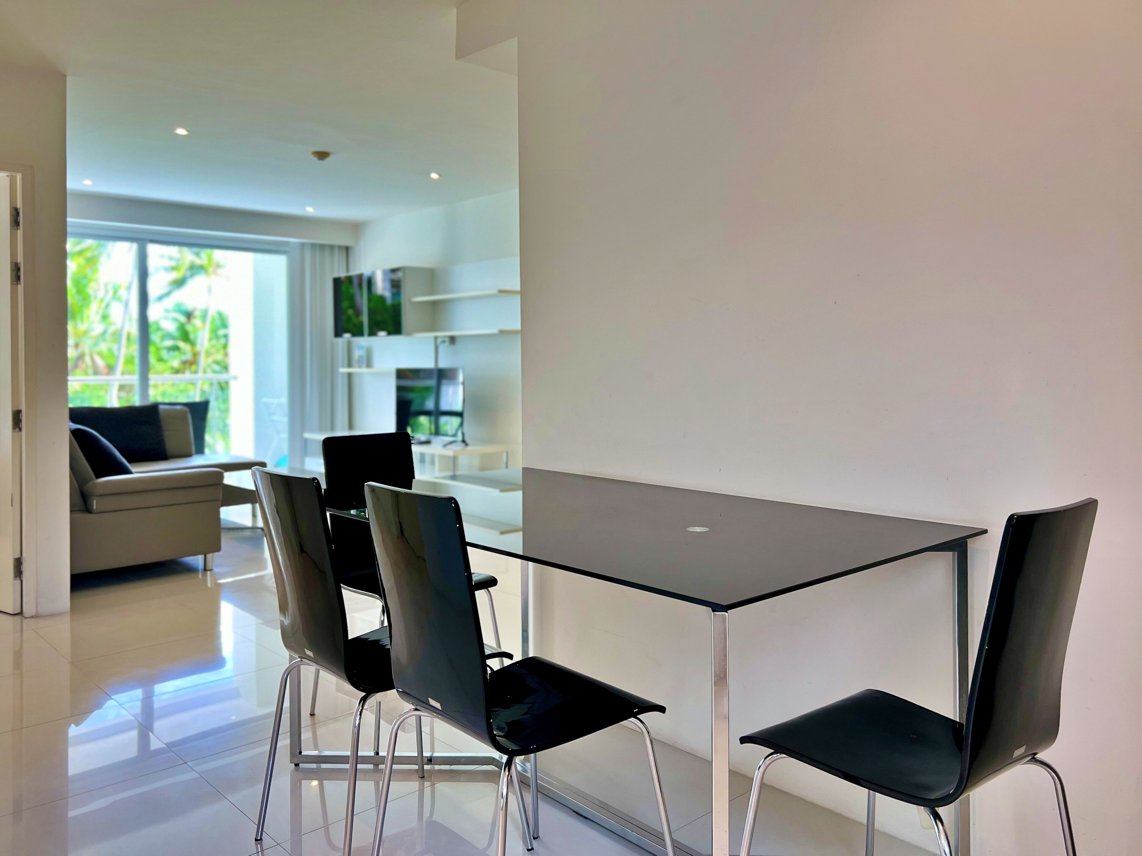 A modern dining area with a glass table and black chairs, a sofa with a view of greenery, and a kitchen area in the background.