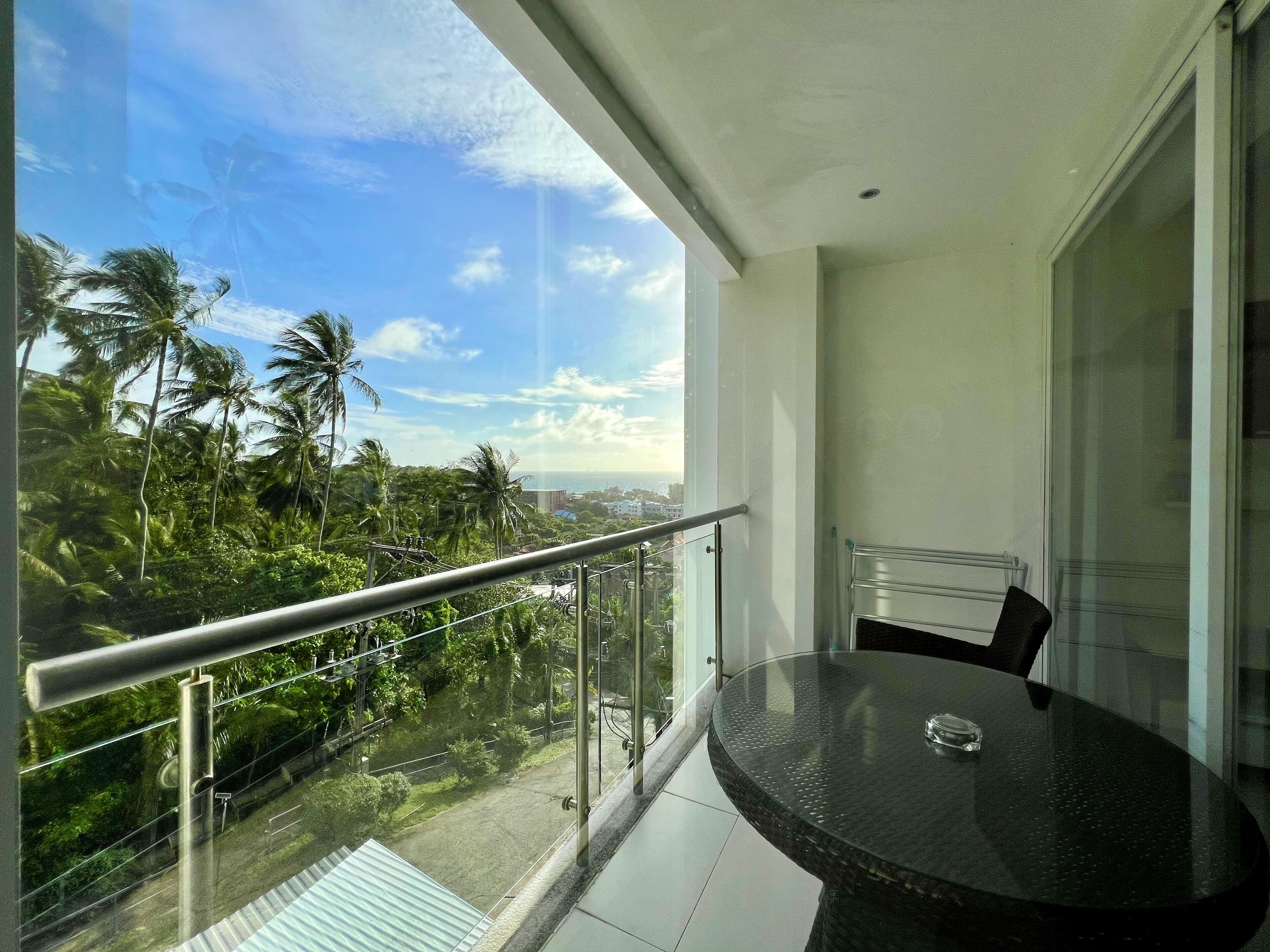 A balcony with a glass railing, a round table, and chairs, overlooking a lush green landscape with palm trees and a clear sky.