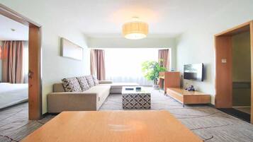 Una habitación de hotel con cama, sofá, mesa de centro, televisor y un amplio ventanal con cortinas.