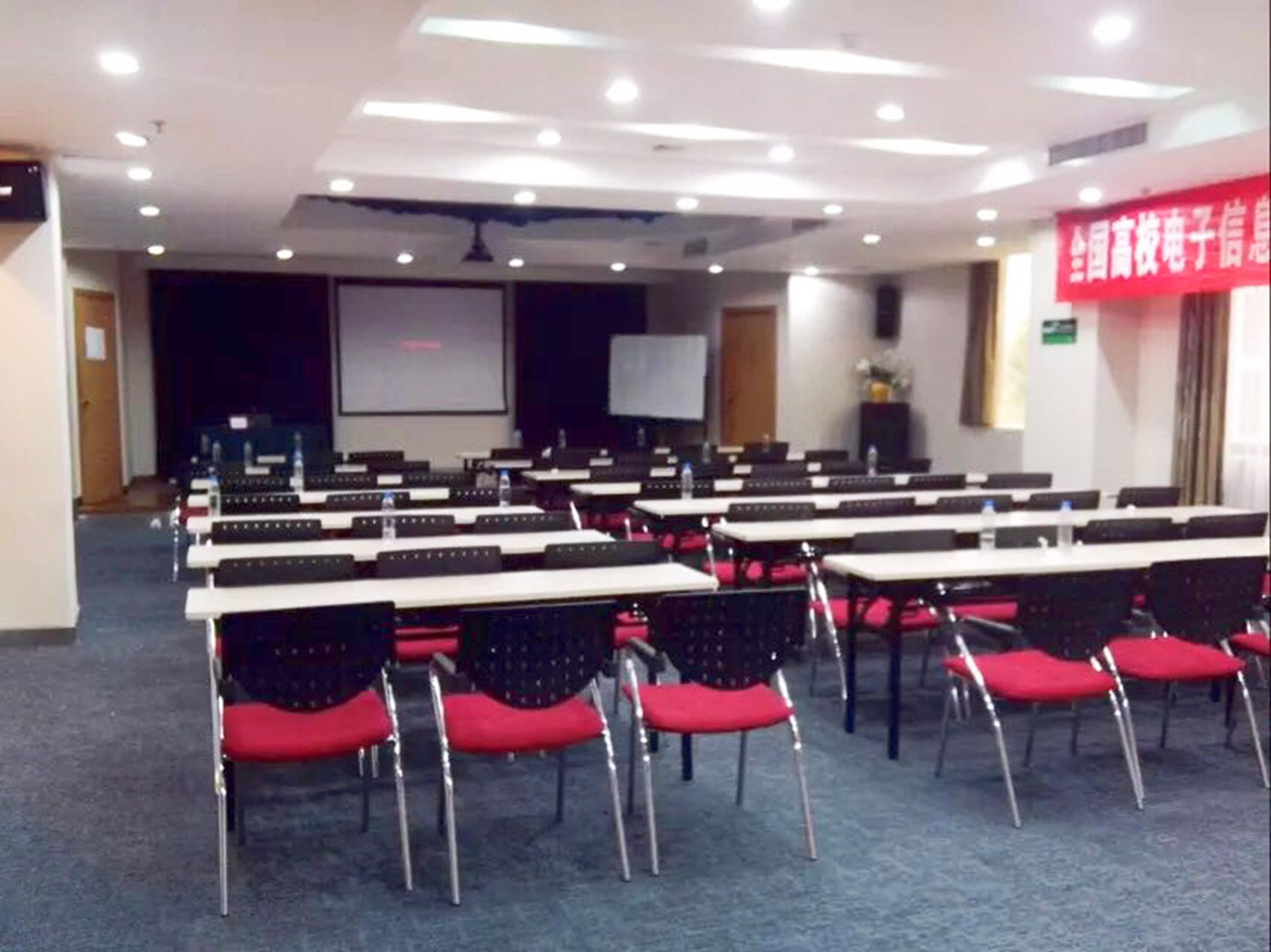 Un salón de conferencias con filas de mesas y sillas, una pantalla de proyector y un estandarte rojo en la pared.