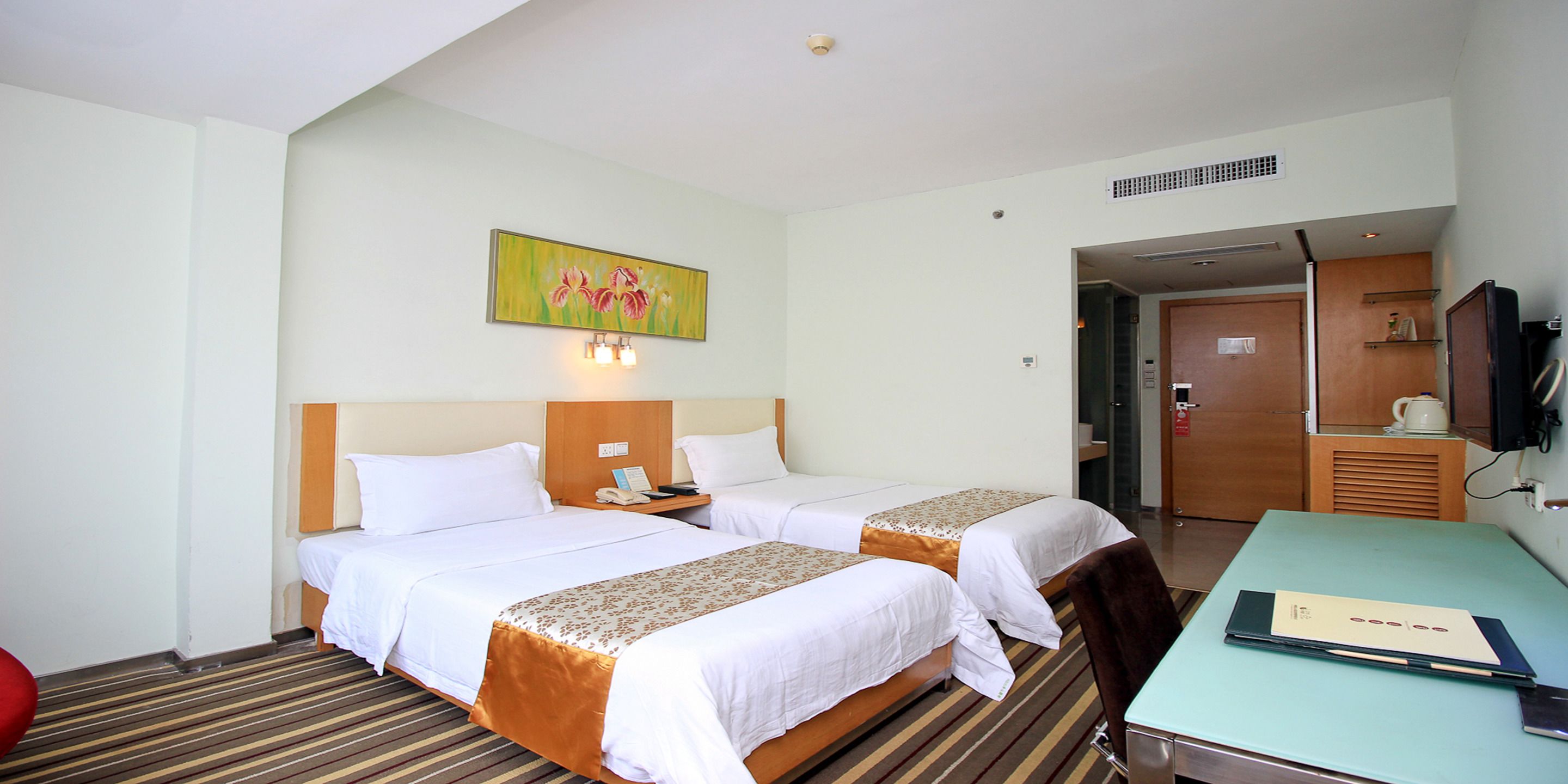 A hotel room with two beds, a desk, a chair, a painting on the wall, and a door leading to a bathroom.