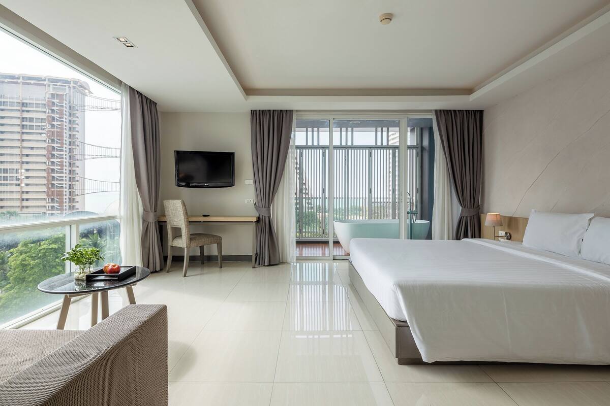 A hotel room with a bed, a desk, a chair, a TV, and a balcony with a view of the city.