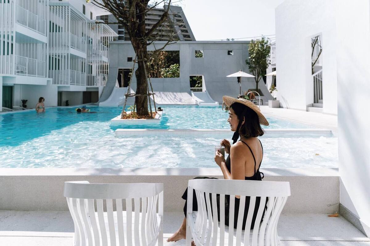 A pool area with a tree, white chairs, and a person sitting by the pool.