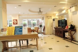 Un hall d’hôtel comprenant un comptoir d’accueil, un lustre, un téléviseur et un panneau indiquant les services proposés, tels que l’organisation d’excursions et la réservation d’hôtels.
