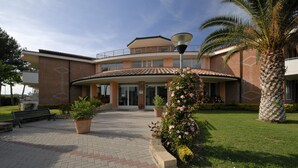 A modern building with a curved facade, large windows, and a central entrance. There are potted plants and a bench in the foreground. - La Cecinella (Cecina)