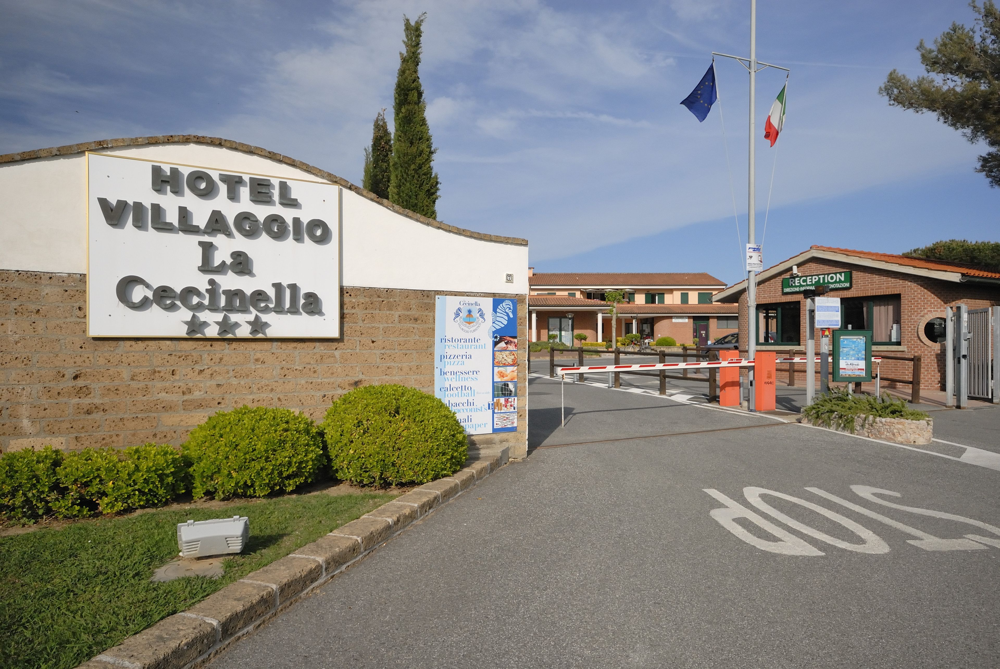 A hotel entrance with a sign for Hotel Villaggio La Cicinella, a reception area, and a flagpole with flags.