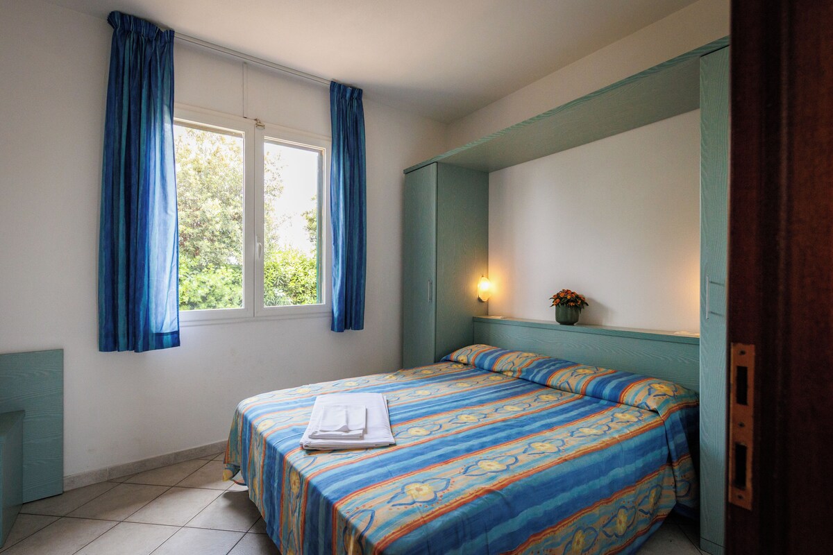 A bedroom with a bed, curtains, a nightstand, a wall-mounted light, and a window with a view of greenery.