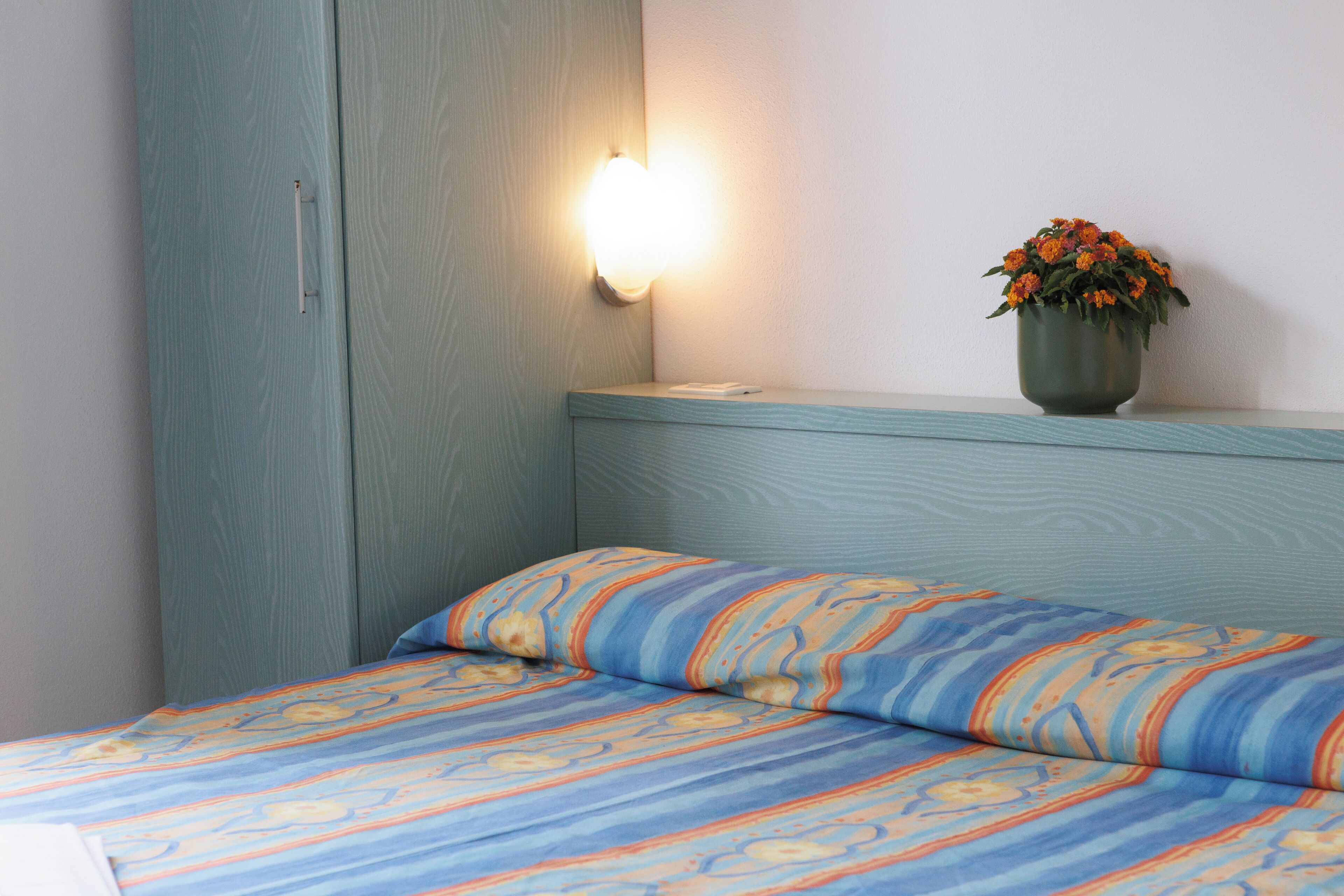 Una cama con una colcha estampada, una mesita de noche con un florero y una lámpara de pared.
