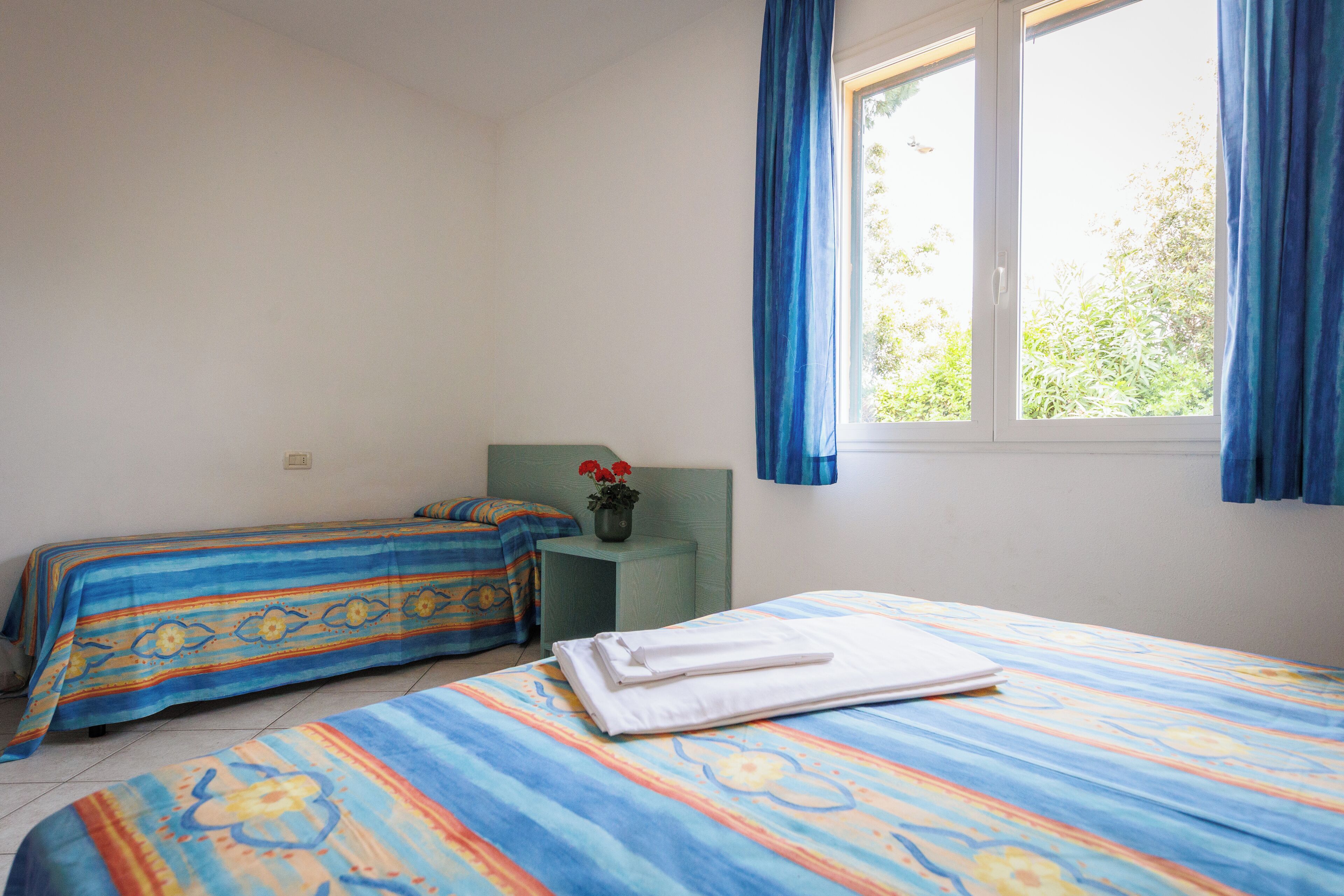 Ein Zimmer mit zwei Betten, einem kleinen Tisch mit einer Vase und einem Fenster mit Vorhängen.