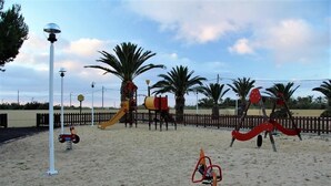 A playground with various play equipment, including swings and a slide, surrounded by palm trees and a fence. - INATEL Porto Santo Hotel (Porto Santo)