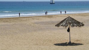 A beach with a thatched umbrella, a sailboat in the distance, and people walking along the shore. - INATEL Porto Santo Hotel (Porto Santo)
